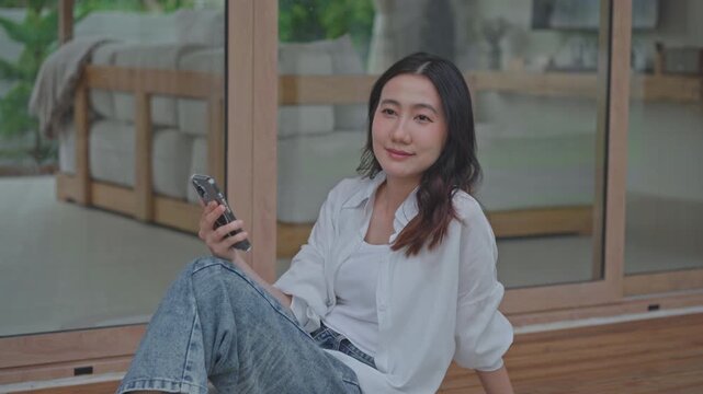 Asian woman sitting on wooden floor holding smartphone, taking a calm break at modern home, enjoying a peaceful mindful moment of slow living and self care