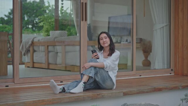 Asian woman sitting on wooden terrace of modern minimal home holding smartphone, relaxing on a calm weekend, enjoying a slow living moment in a cozy living space
