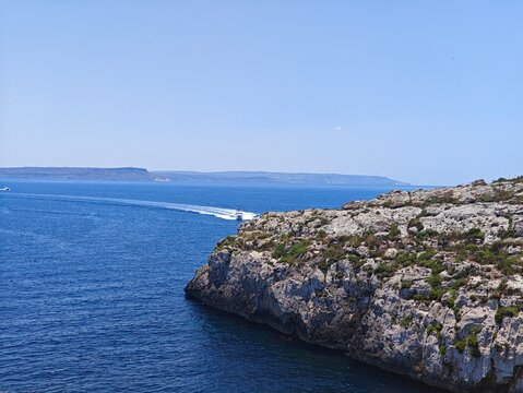 Rocky coastline on Gozo, Malta, overlooking the deep blue Mediterranean Sea with a speeding motorboat leaving a white wake under a clear summer sky