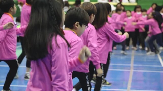 사람 스포츠 훈련 코트 야외 일상 소품 디테일 자재: 실내 체육관 푸른색 코트 바닥 위에서 분홍색 단체복 상의를 입고 대형을 유지하며 매스게임 연습 동작을 수행하는 사람들의 옆모습과 움직임을 포착한 미디엄 전경 동영상 클립입니다.