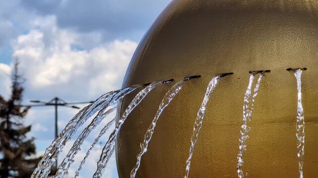 Close-up of a spherical golden fountain featuring multiple streams of water gracefully flowing out. Background consists of a cloudy sky and blurred trees, creating a serene outdoor atmosphere.
