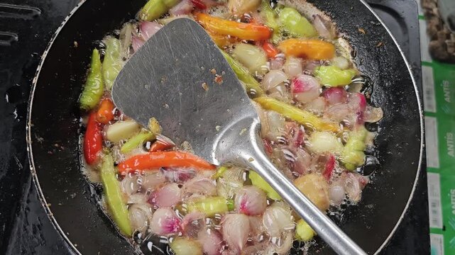 Frying fresh whole shallots and colorful hot chili peppers in bubbling oil with a metal spatula inside a black pan