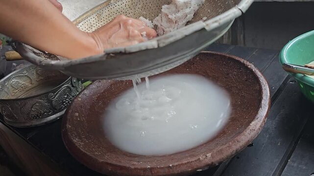 A person manually squeezing grated coconut in a metal sieve to extract fresh coconut milk into a traditional clay bowl
