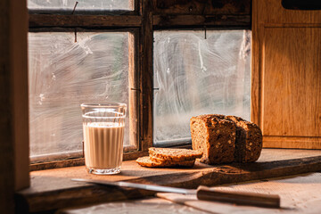 Freshly baked bread with seeds on a rustic wooden board by an antique window, next to it stands a glass of a traditional drink, along with slices of bread and knife, all bathed in warm, natural light © Владимир Трубачёв