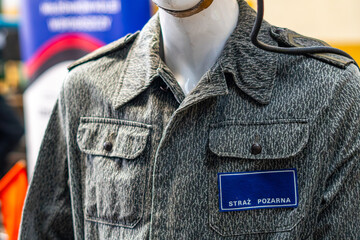 Close-up of a fire brigade uniform in grey camouflage pattern worn on a mannequin, featuring a dark blue ID patch with the text STRAŻ POŻARNA, chest pockets, buttons, and epaulets in an industrial int © Adam