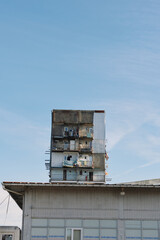 Naklejka premium Tall stacked apartment tower atop a low industrial building under a clear blue sky, with small figures on balconies, urban housing structure and exposed living spaces.