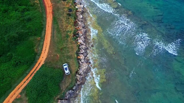 The aerial view captures a winding seaside road bordered by lush greenery, where a lone car drives along the rocky coastline, waves crashing against the shore.