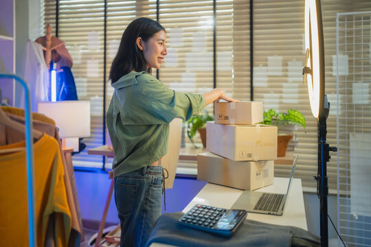 Asian woman live streaming unboxing and packing clothing product in home office, online retail e-commerce entrepreneur, small business owner logistics fulfillment and social commerce concept