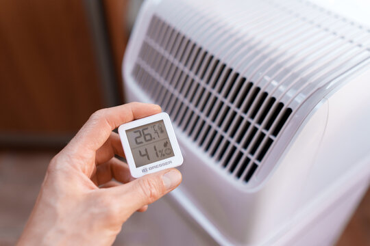 Close-up of a hand holding a room thermometer showing temperature in front of a portable air conditioner