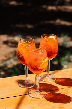 Three glasses of refreshing Aperol cocktail drinks sit on a wooden table in a sunny outdoor setting in a garden during the afternoon