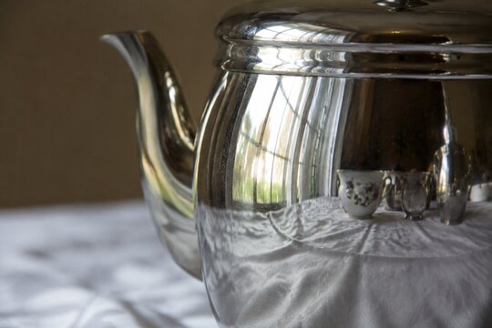 A silver coffee pot reflecting a distorted window and tablecloth.