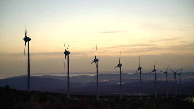 A row of wind turbines stands on a hillside as the sun sets behind them, generating clean energy. The turbines' white blades rotate in the breeze, surrounded by sparse vegetation.