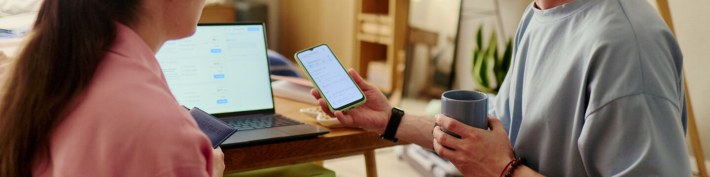 Header of young adult couple preparing for vacation, checking smartphone booking details beside laptop and passport, planning travel logistics. Ideal for tourism marketing, trip planning apps