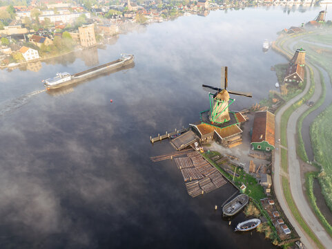 Aerial view of Zaanse Schans windmills along the Zaan river with a cargo barge navigating the misty water in Zaandam, Noord-Holland, Netherlands.