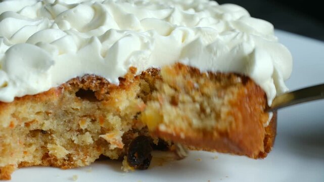 Close up golden spoon taking piece of moist homemade carrot cake topped with swirls of delicious cream cheese frosting, revealing texture with raisins and grated carrots. Person eating carrot cake 