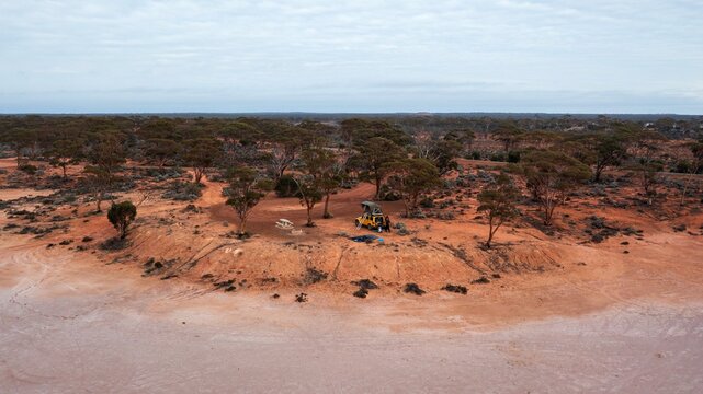 Aerial view of a yellow off-road vehicle with a rooftop tent parked on red earth near sparse trees in the arid outback of Norseman, Western Australia, Australia.