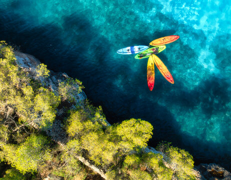 Aerial drone view of colorful kayaks in crystal clear turquoise sea near rocky coast and green trees. Summer outdoor adventure in Mallorca, Balearic Islands, Spain. Top down view of canoes. Travel