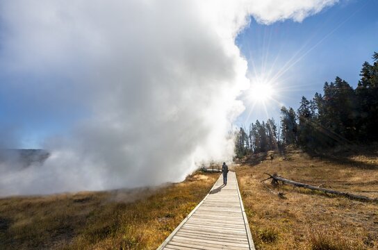 Hiker on footbridge next to steaming hot spring, sun with sun star, Churning Caldron, Yellowstone National Park, Wyoming, USA