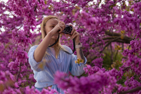 Young woman standing amidst blooming purple flowers, holding a retro camera and capturing the beauty of a flowering tree in spring, engaged in her photography hobby