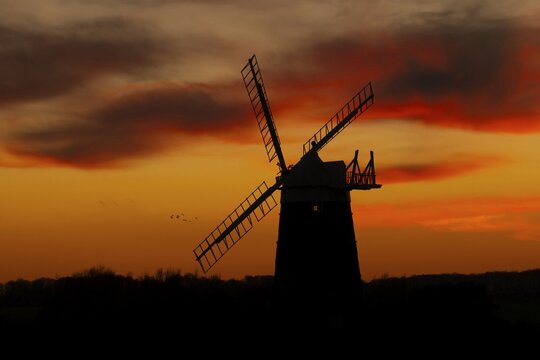 Windmill silhouette at sunset with a red sky and clouds and a flock of Lapwing flying behind, Burnham Ovary Staithe, Norfolk, England, United Kingdom