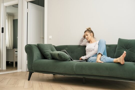 Middle-aged woman sits comfortably on a green couch in a stylish living room, focused on her laptop. The airy space features soft lighting and minimalist decor, creating a calming atmosphere
