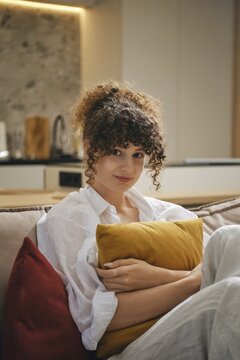 A young woman with curly hair sits comfortably on a couch in a stylish living room. She hugs a yellow pillow and wears a casual outfit, appearing relaxed and content in her serene environment
