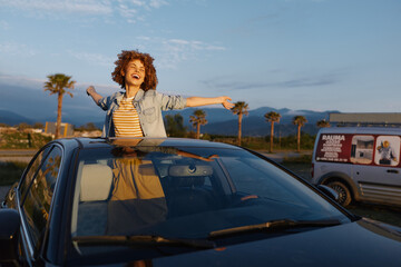Fototapeta premium Joyful young woman leans from a car sunroof with arms wide in a sunlit coastal lot, capturing carefree travel vibes.