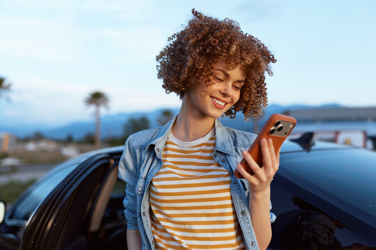 Young woman with curly hair smiles while checking her phone beside a car on a sunny day