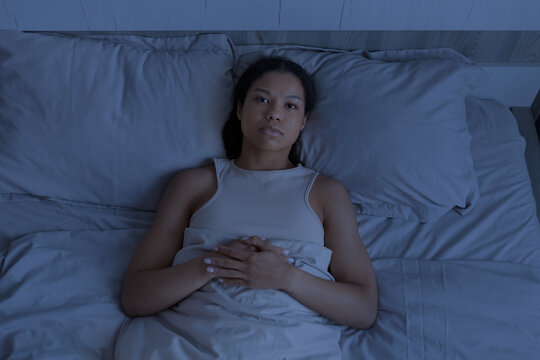 Black woman lying awake in bed at night with serious expression, suggesting insomnia, worry, loneliness and quiet reflection in a dark bedroom