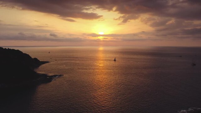 Bright orange sun setting over the Andaman Sea, golden reflections on the water with a single sailboat passing along the scenic coastline of Nai Harn Beach in Phuket, Thailand. Aerial view drone shot