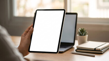 Fototapeta premium A woman's hand holding a white screen tablet, with a laptop and notebooks on a wooden desk in the background, perfect for showcasing digital content or mockups