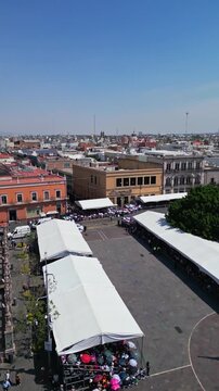 Aerial drone flight over Plaza Patria with the fair market tents and a distant crowd in downtown Aguascalientes on a sunny day, central Mexico.