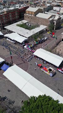 Aerial drone flight over Plaza Patria with fair tents and people gathering in Aguascalientes on a sunny day, central Mexico.