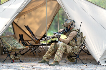 Fototapeta premium One army man or soldier sit on chair and check his weapon in front of military tent with daylight