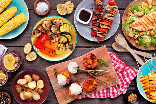 Summer BBQ food table scene over a dark wood background. Grilled meat skewers, corn, potatoes, vegetables, pineapple, peaches and ice cream, salad and iced tea. Top view.