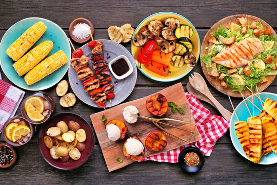 Summer BBQ food table scene over a dark wood background. Grilled meat skewers, corn, potatoes, vegetables, pineapple, peaches and ice cream, salad and iced tea. Overhead view.