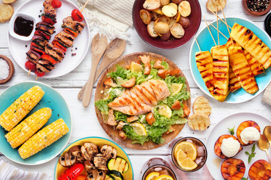 Summer BBQ food table scene over a white wood background. Grilled meat skewers, corn, potatoes, vegetables, pineapple, peaches and ice cream, salad and iced tea. Overhead view.