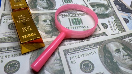Gold bar and pink magnifying glass on U.S. currency, symbolizing financial analysis, wealth evaluation, and investment © MR HEPPY