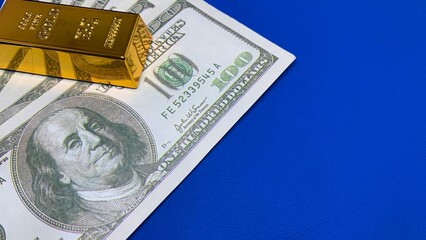 Gold bar positioned on U.S. hundred-dollar bill against blue background, symbolizing investment value and financial security © MR HEPPY