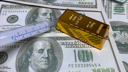 Plastic syringe beside gold bar on U.S. hundred-dollar bills, representing healthcare costs and financial investment relationships © MR HEPPY