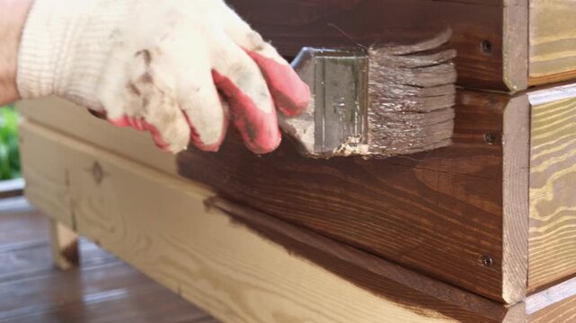 Brown oil for protection of wood by staining. Painting fence for protecting and pests. Woodworker applying finish to pine board box. Repairman varnishing with brush wooden fencing wall plank surface.