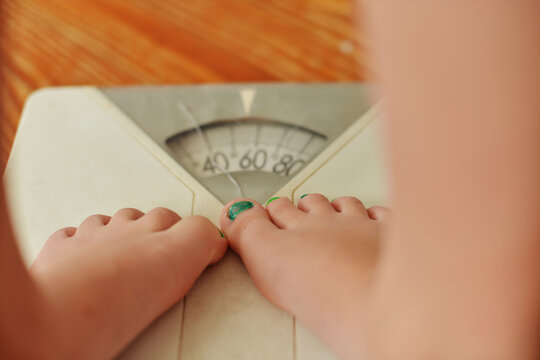 Cracked analog bathroom scale beneath a young girl's bare feet with green painted toenails, viewed from above to illustrate weight measurement, childhood growth, wellness, and personal health.