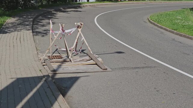 Wooden sawhorses and caution tape marking an open sewer manhole on the side of a street during road repair or maintenance work