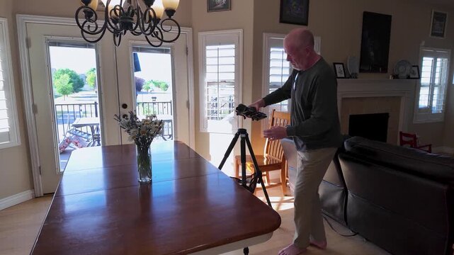 A bald senior man setting up a camera slider and tripod in a sunny living room for a video production shoot