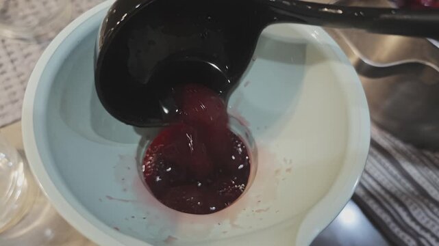 Close up top view of a person ladling cooked plum preserves through a funnel into a glass jar, showing the process of making jam at home