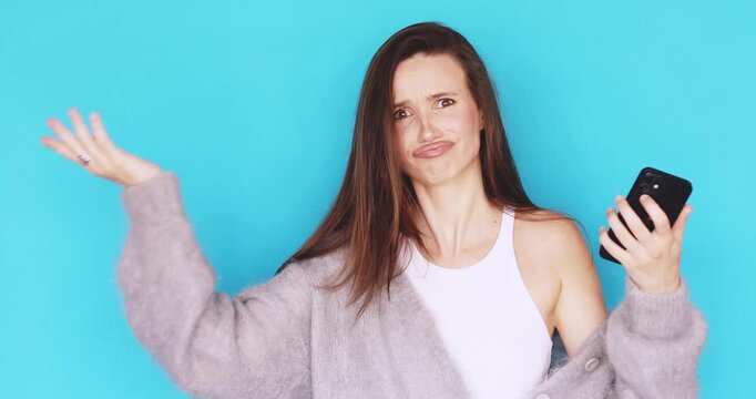 Young woman looking at smartphone with annoyance and frustration, expressive face showing disbelief, gesturing with hands, studio portrait isolated on blue background, concept of technology problems