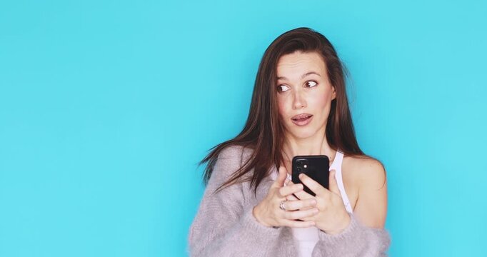 Young woman looking at smartphone screen with skeptical and puzzled facial expression, confused brunette girl browsing mobile phone with raised eyebrow, studio isolated on blue background, tech issue