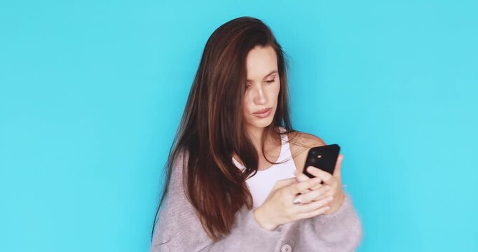 Young woman looking at smartphone screen with neutral and pensive facial expression, brunette girl browsing mobile phone with serious look, studio portrait on blue background, tech use, concentration