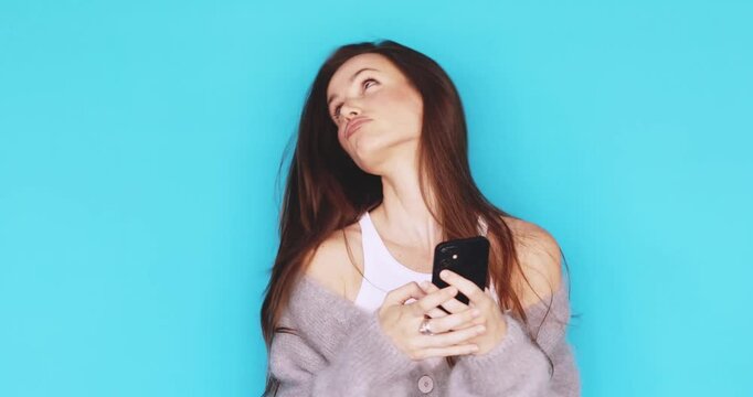 Young woman looking at smartphone with skeptical and puzzled facial expression, confused brunette girl browsing mobile phone with raised eyebrow, studio isolated on blue background, tech issue, doubt