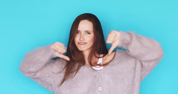 Young woman showing thumbs down gesture with skeptical and disappointed facial expression, brunette girl gesturing dislike, disapproval and negative reaction, isolated on blue background, rejection